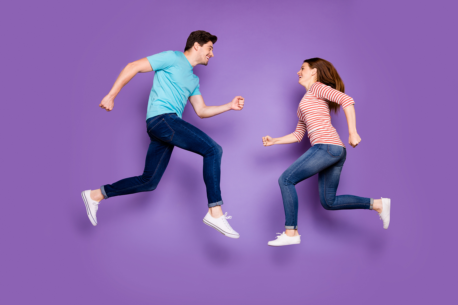 A man and a woman wearing blue and pink against a purple background, jumping high in the air looking at eachother and smiling.