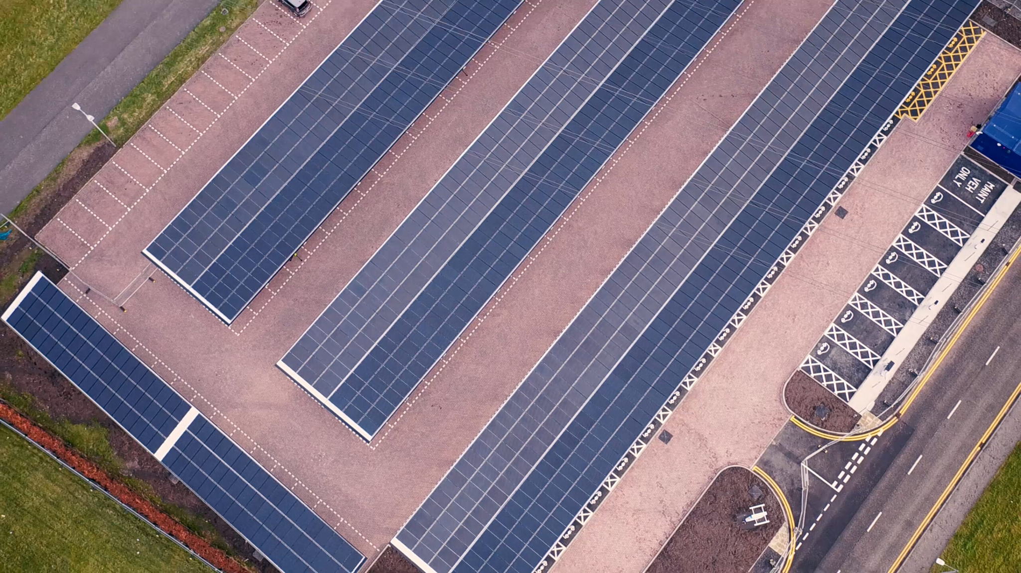 Aerial view of large solar panel arrays on a flat rooftop. The panels are aligned in neat rows across a concrete surface. Adjacent areas include roads and green landscaped sections. No people or vehicles are visible.