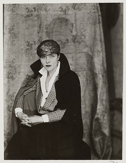 Black and white image of Djuna wearing several layers sitting casually in a chair with a serious expression.