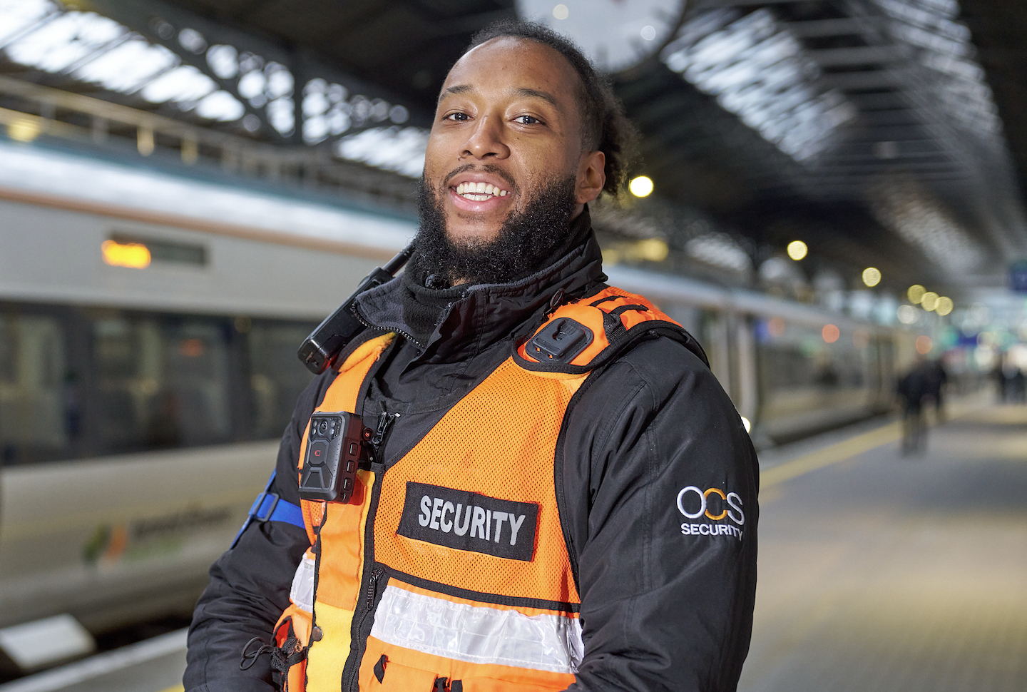 A smiling security guard wearing an orange vest and black jacket stands on a train station platform with a train in the background.