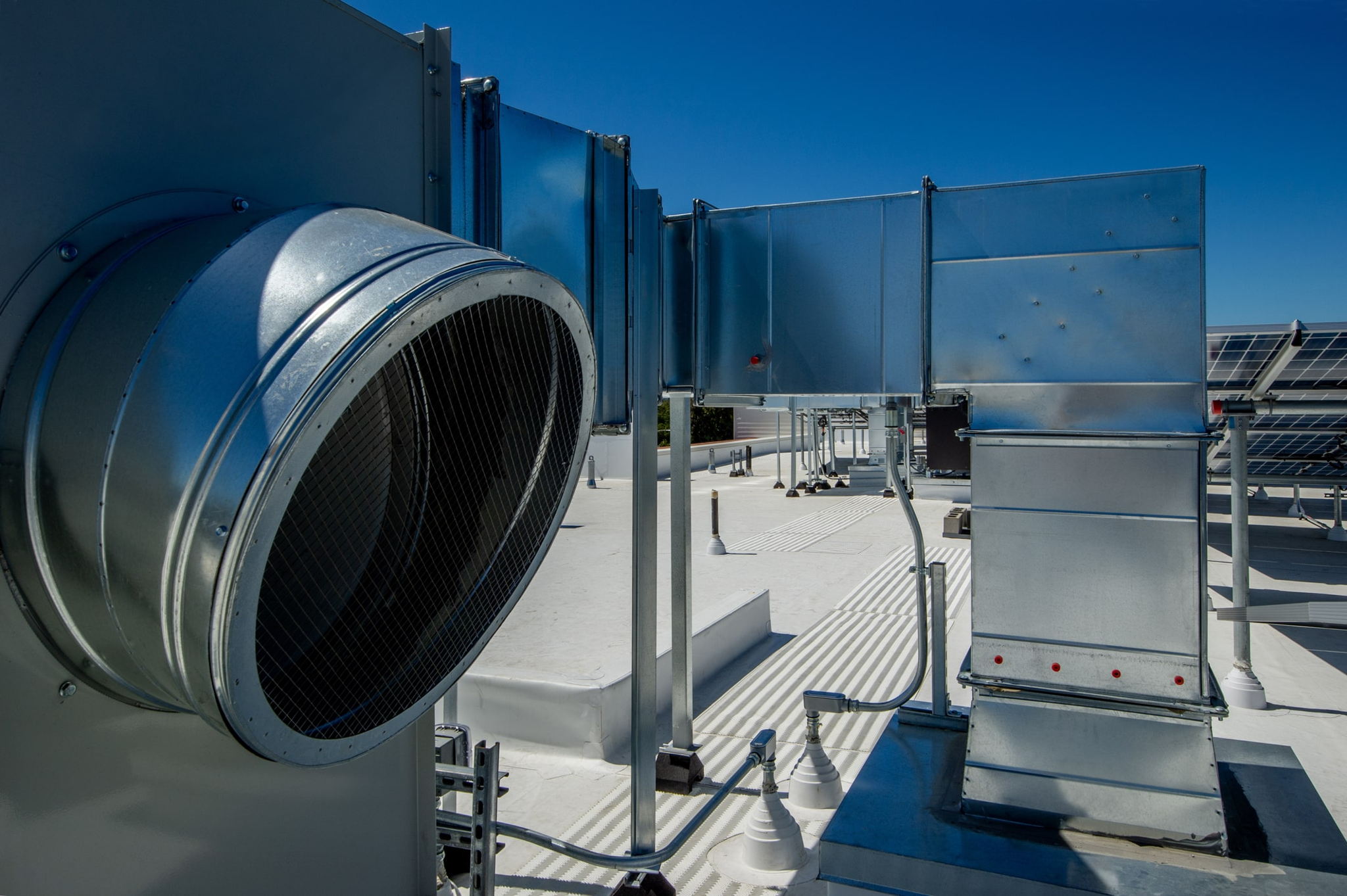 Large industrial HVAC ductwork and ventilation system on a white rooftop, with metal pipes and panels under a clear blue sky.