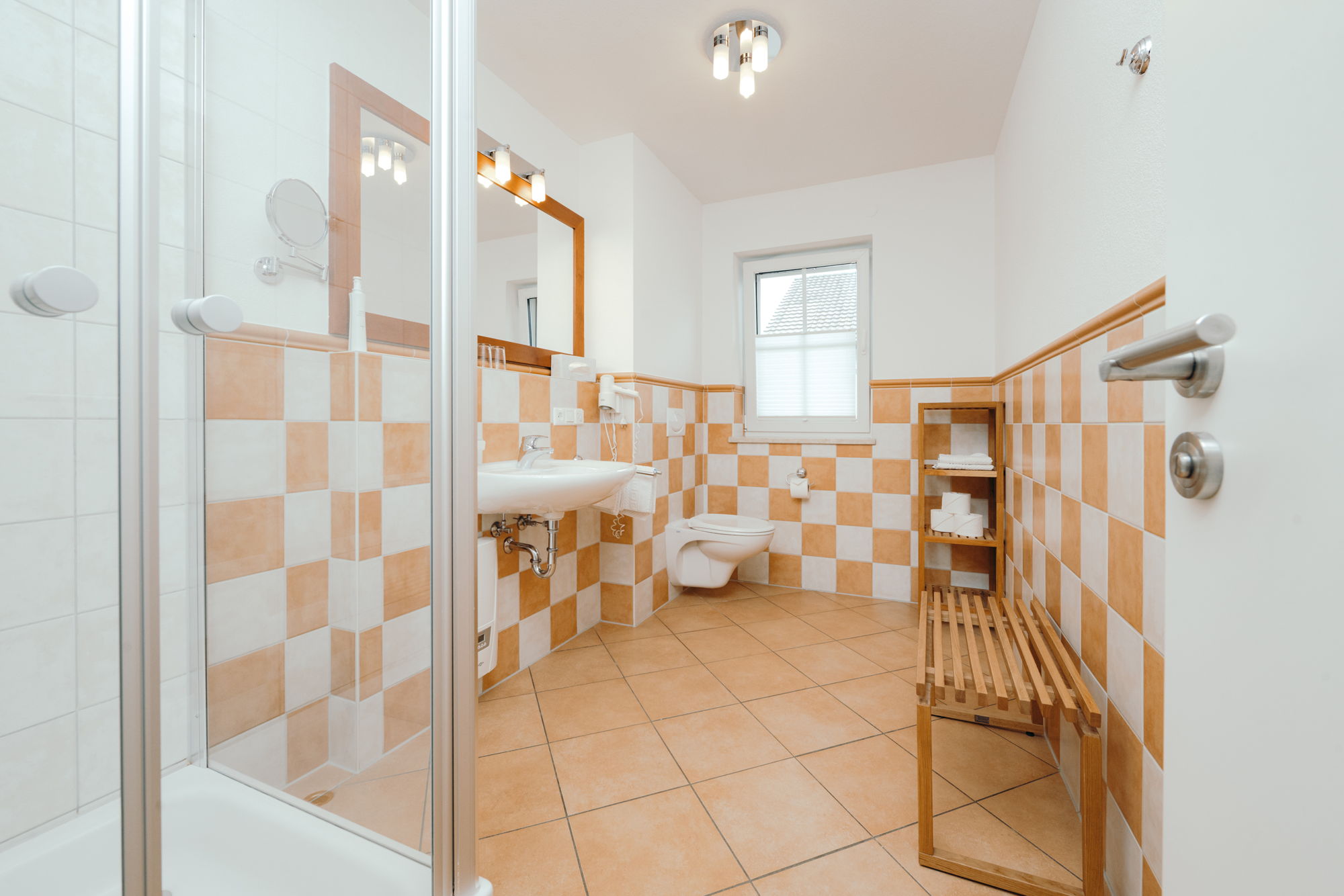 Ein Badezimmer mit orangenem Fliesenboden, weiß-orange karierten Wandfliesen, Dusche, Waschbecken, Toilette, Fenster und einer Sitzbank.