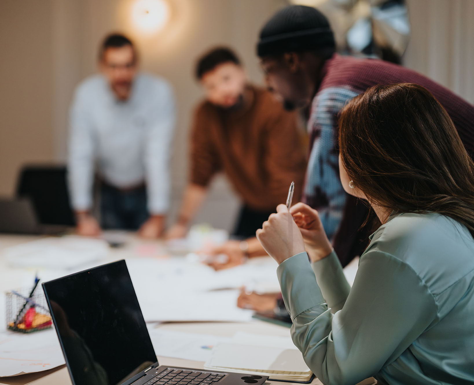 Four people gather around a table covered with papers and a laptop, engaged in discussion. The focus is on a woman holding a pen, while three others stand slightly out of focus in the background.