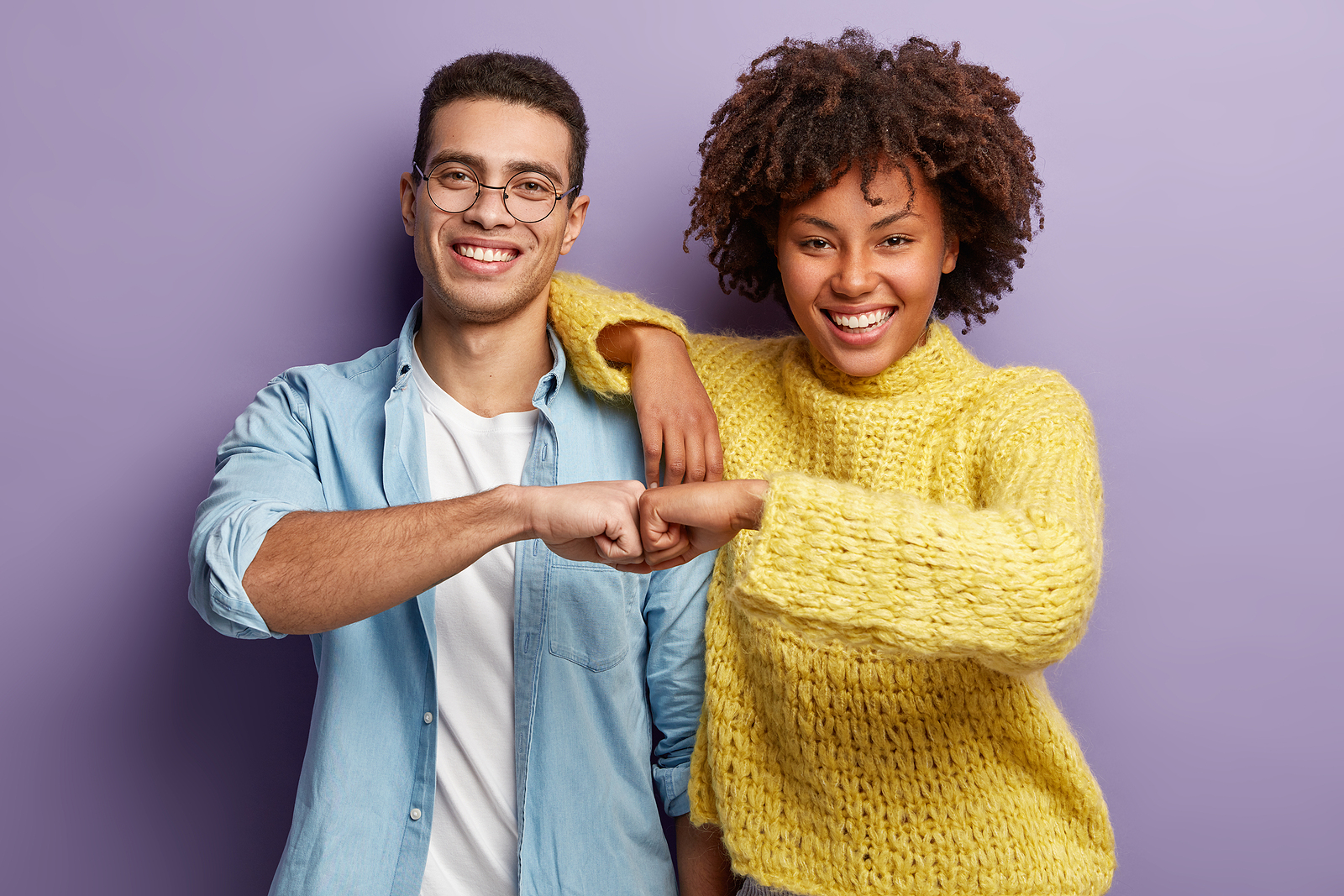 An attractive man and woman of different ethnicities fist bumping and smiling wearing comfortable clothes against a purple background.