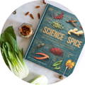 A book, The Science of Spice, lays on a table surrounded by loose spices and vegetables