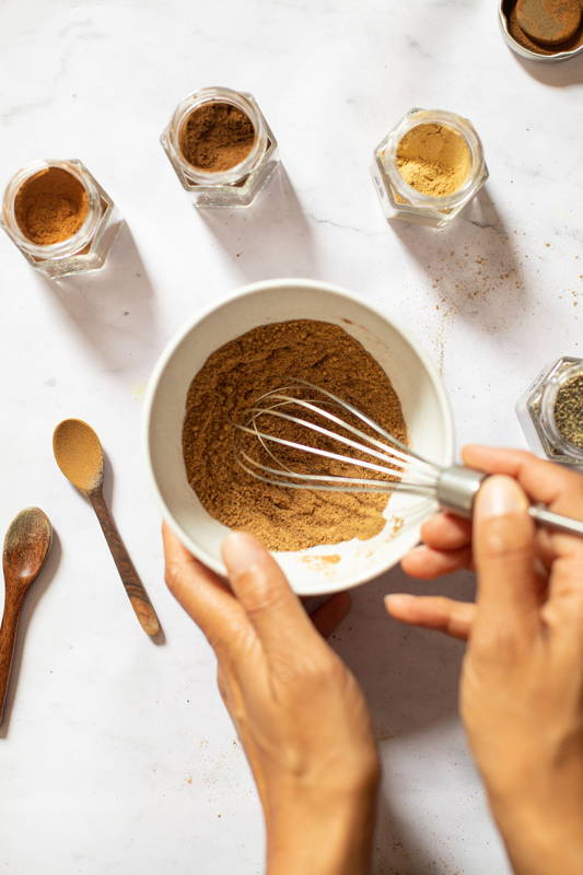 Brownish ground spice is being whisked in a white bowl surrounded by open glass spice jars