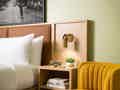 Moderne Zimmerecke mit Kingsize-Bett, Holz-Kopfteil, Wandlampe und einem senfgelben Polstersessel.