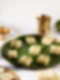 Squares of white Indian barfi topped with crushed pistachios on a banana leaf–lined plate, with a brass lamp and cup in the background.