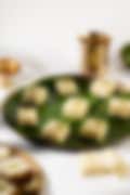 Squares of white Indian barfi topped with crushed pistachios on a banana leaf–lined plate, with a brass lamp and cup in the background.