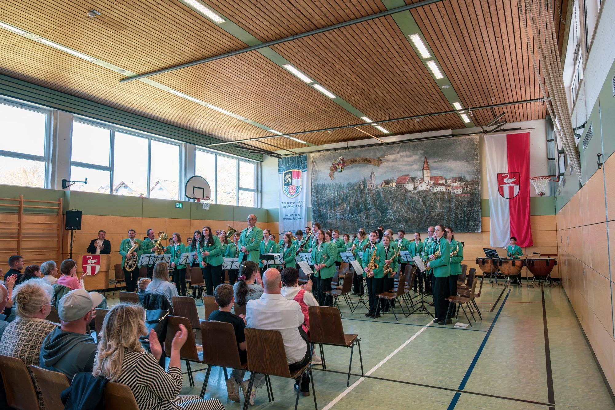 Ein Blasorchester in grüner einheitlicher Tracht steht in einer Turnhalle vor Publikum. Alle halten ihre Blasinstrumente vor sich. 