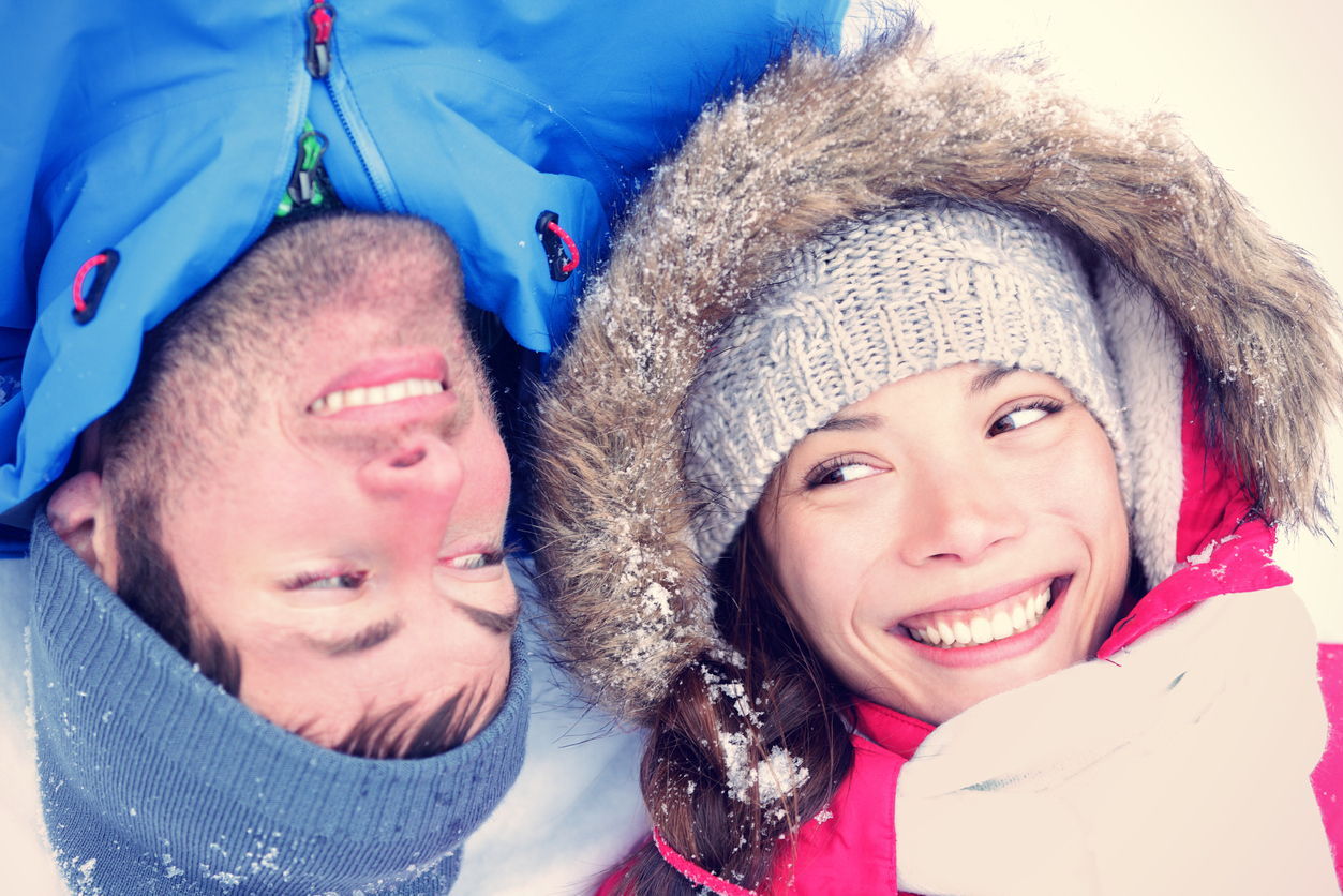 A man and a woman laying on the snow side by side look to eachother and smile.