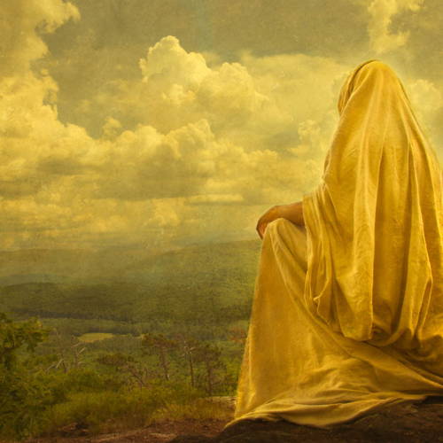 Jesus in a yellow robe sitting on a hill and looking out at the landscape.