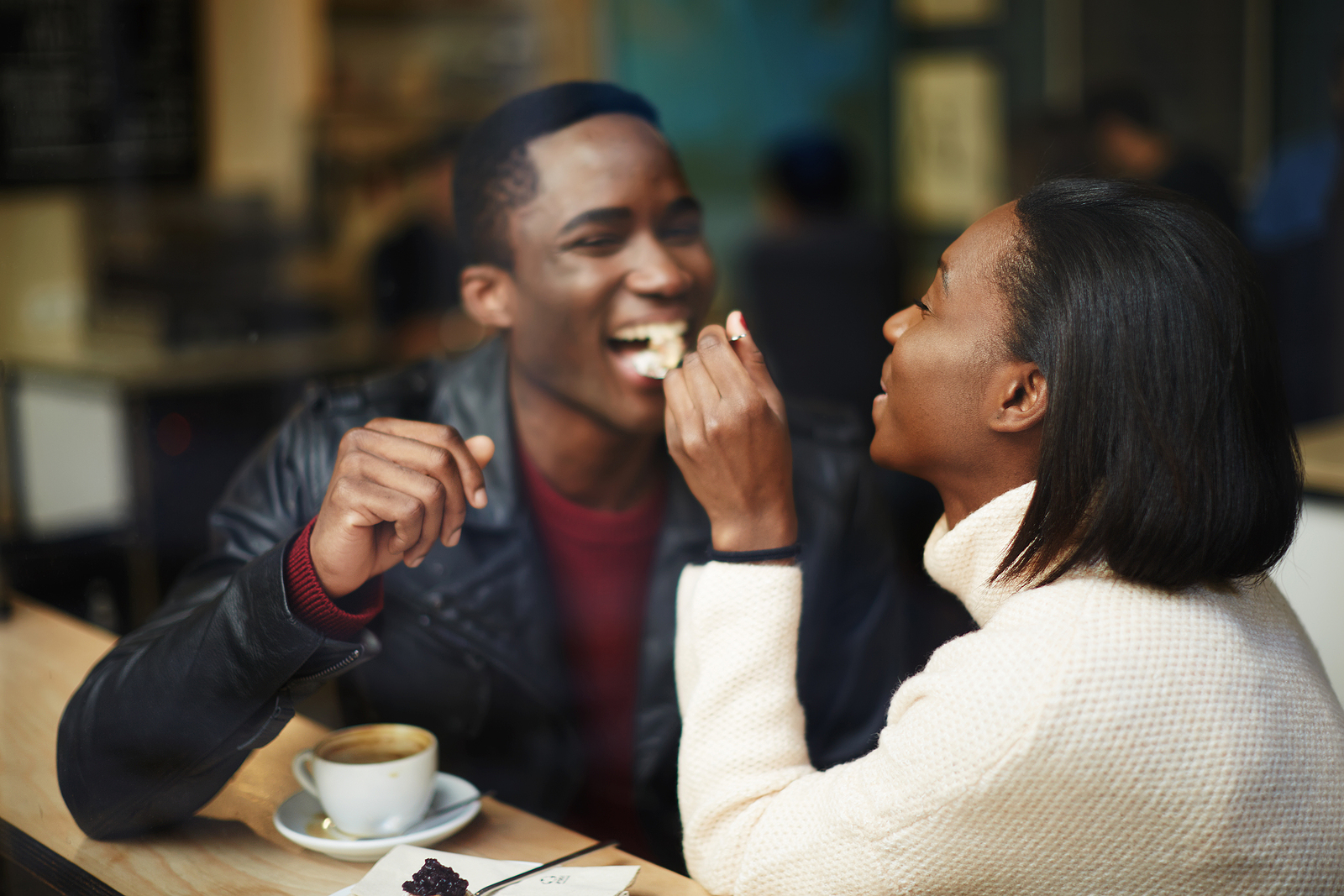 Image of a black young couple at a coffee shop, the woman is giving a spoonfull of food to her friend and he is smiling.