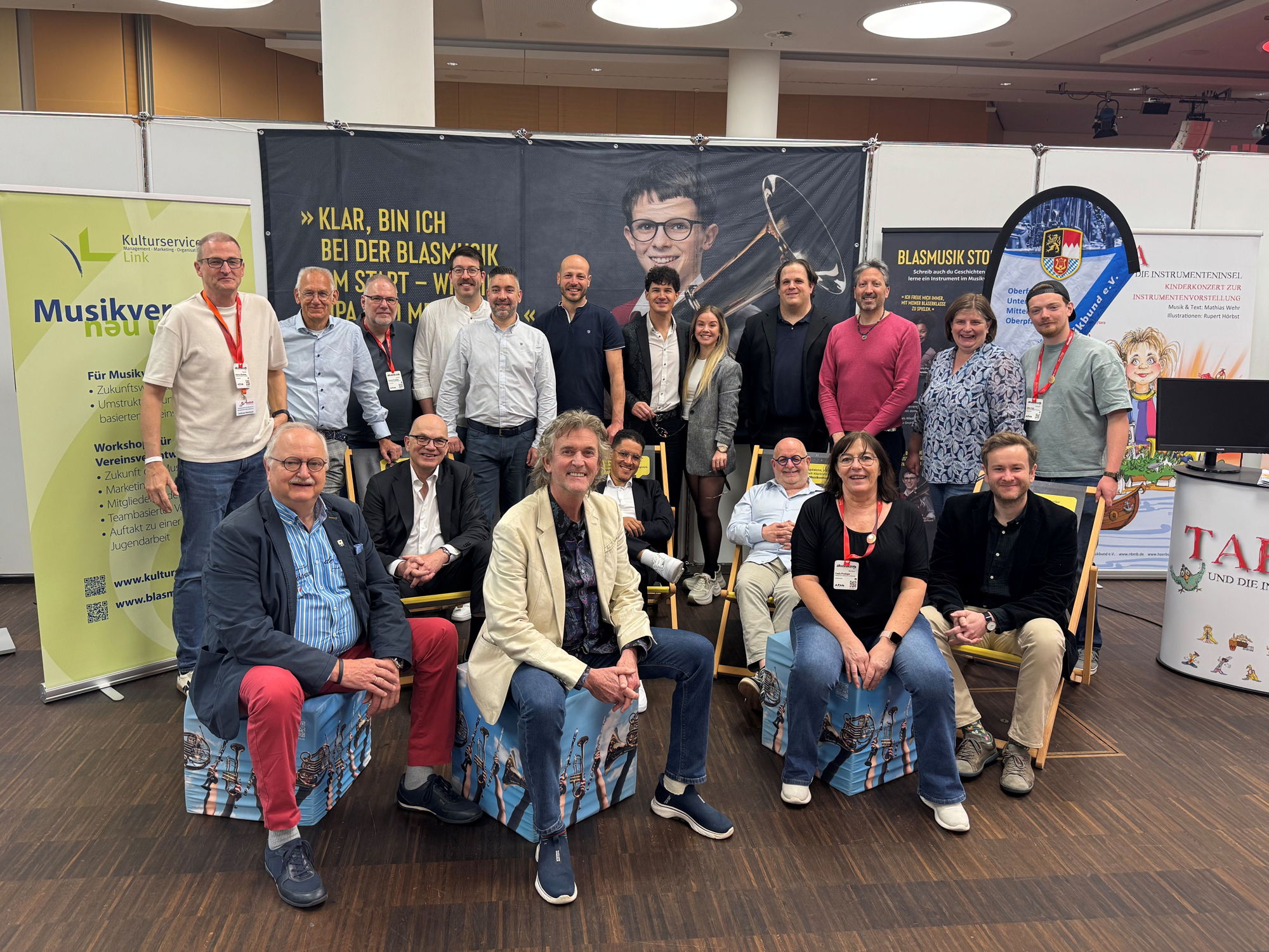 Viele Menschen stehen und sitzen vor einem Messe-Stand und lächeln in die Kamera. 