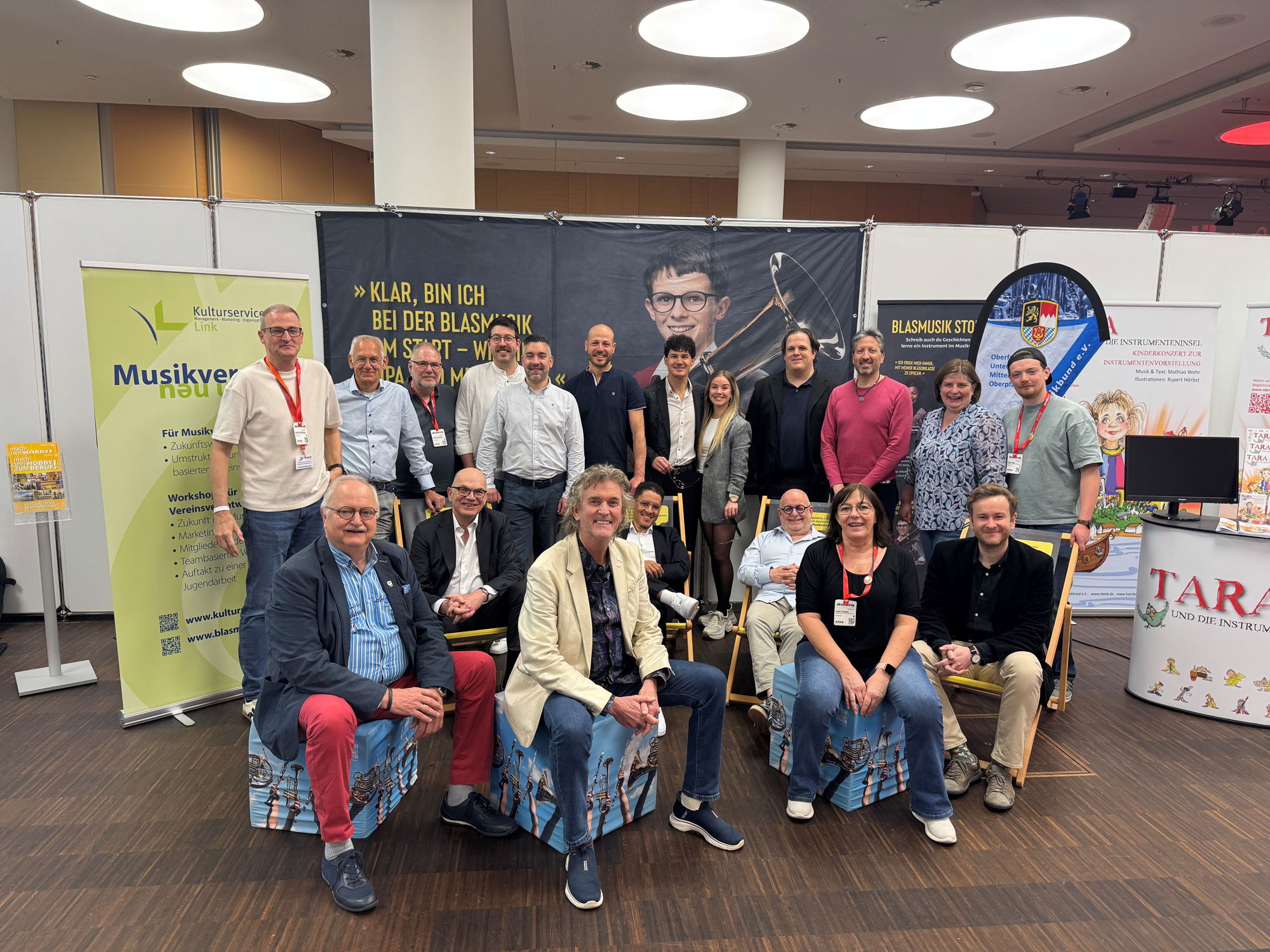 Viele Menschen stehen und sitzen vor einem Messe-Stand und lächeln in die Kamera. 