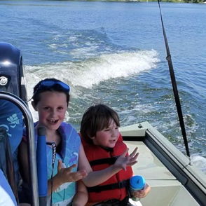 Two joyful children on a boat, one in a life jacket, enjoying a sunny day on the water, capturing playful summer moments.