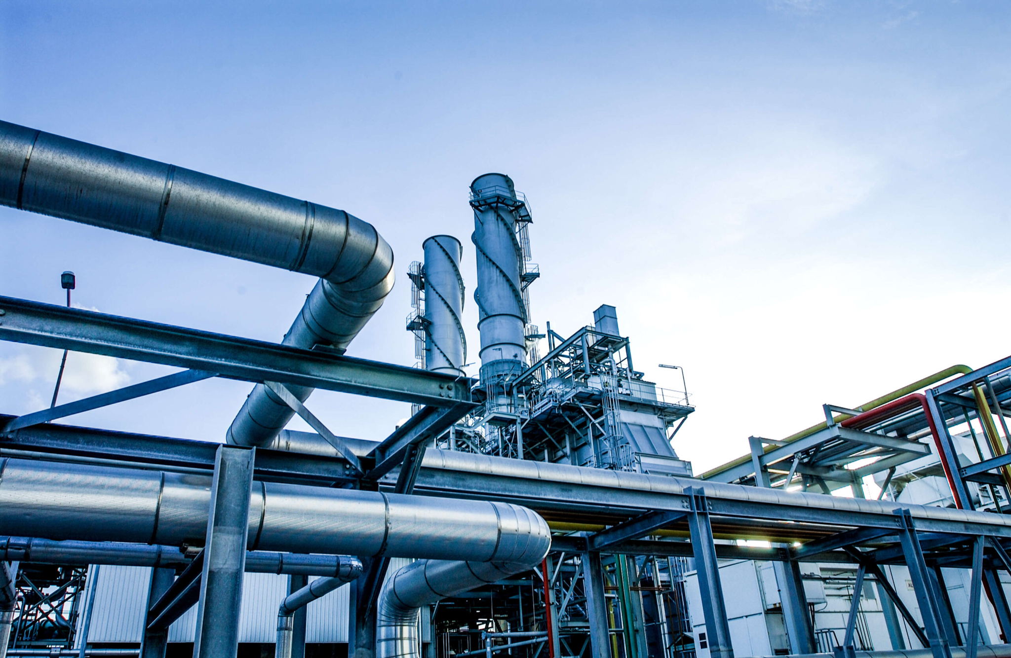 Exterior view of an industrial facility with large metal pipes and chimneys against a blue sky. The structure is composed of complex piping and metal frameworks, showcasing a modern industrial setting under a clear sky.