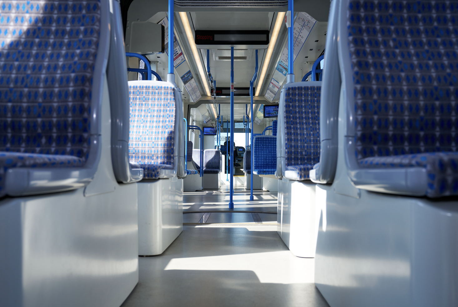 Interior of an empty modern train carriage, featuring blue patterned seats and metal poles, with sunlight streaming in through the windows, viewed from a low angle facing the front of the train.