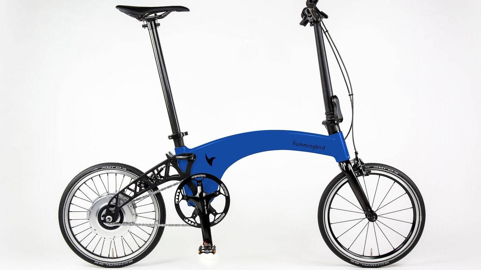 Zijaanzicht van een blauwe elektrische vouwfiets met compact frame, kleine wielen, hoog zadel en stuur rechts, vrijstaand op een neutrale achtergrond.