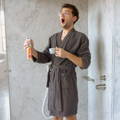 A man wearing a bathrobe stands inside a modern shower, holding a cup of coffee in his left hand and a bathroom spray bottle in the other. He is spraying the glass walls of the shower and appears to be yawning. The shower features sleek, contemporary design elements and is well-lit.