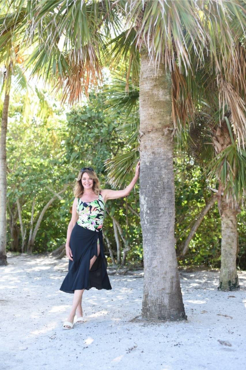 A woman in a rainforest rainbow swimsuit with a black sarong lounges next to a palm on the beach.