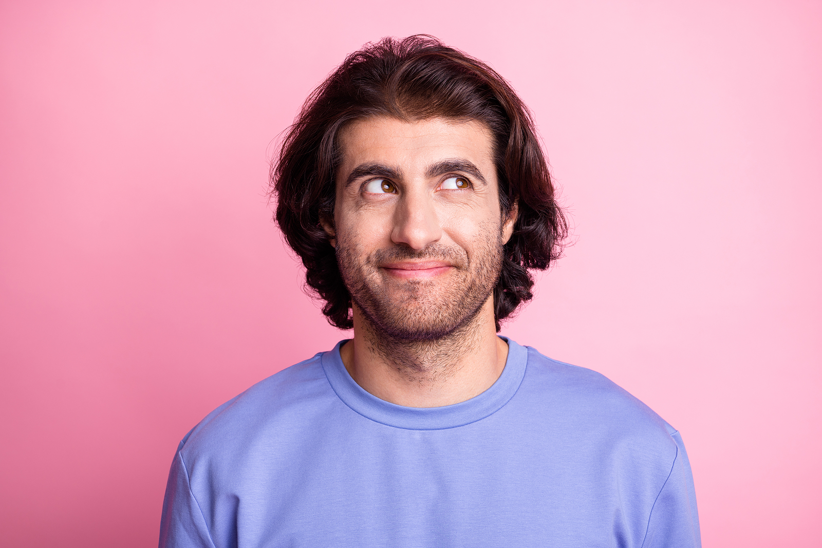 An attractive man smiles while looking to his side against a pink background.