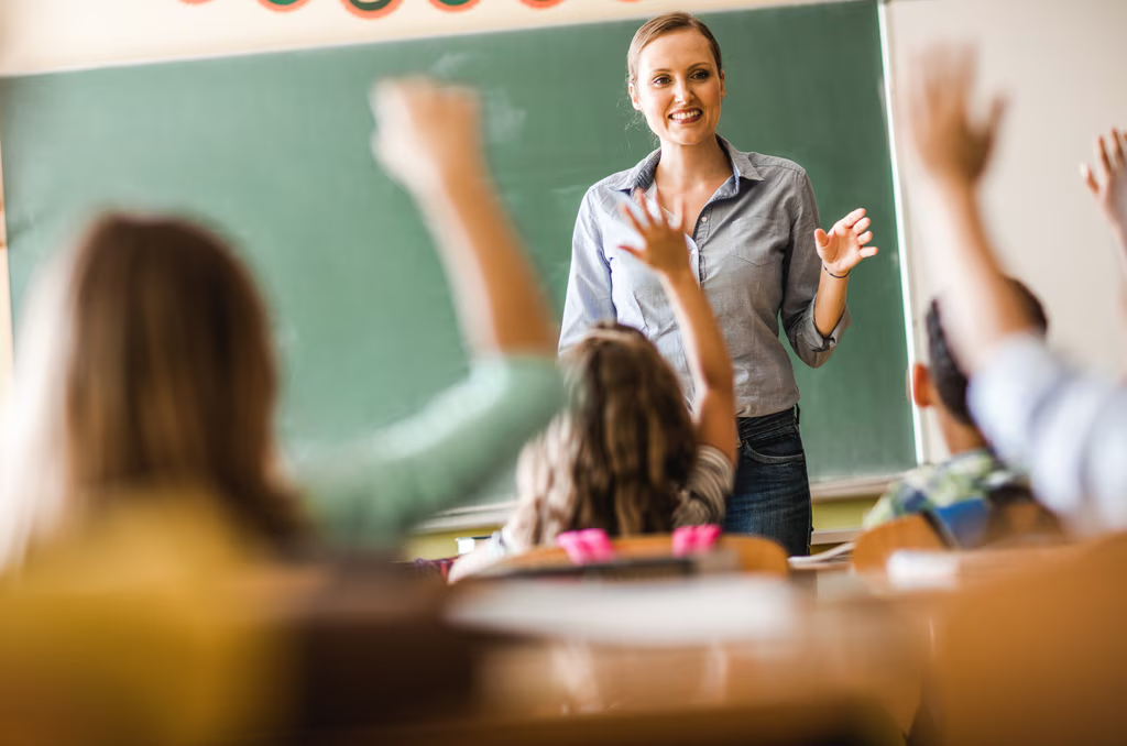 A teacher stands and smiles in front of a chalkboard while several students, seen from behind, raise their hands in a classroom.
