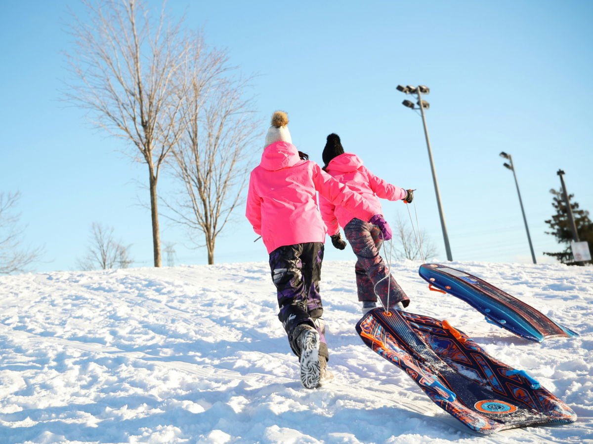 two children in ski suits race up a hill with a toboggan