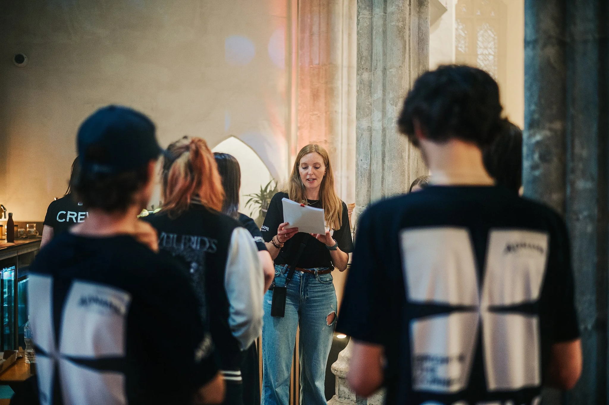 A woman stands in front of a small group, reading from a clipboard in a softly lit room, while several people wearing matching T-shirts with large white squares on the back listen attentively.