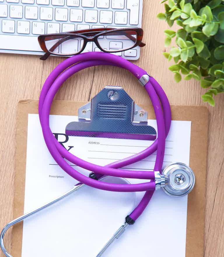 Purple stethoscope on a clipboard with a medical form, glasses, keyboard, and plant.