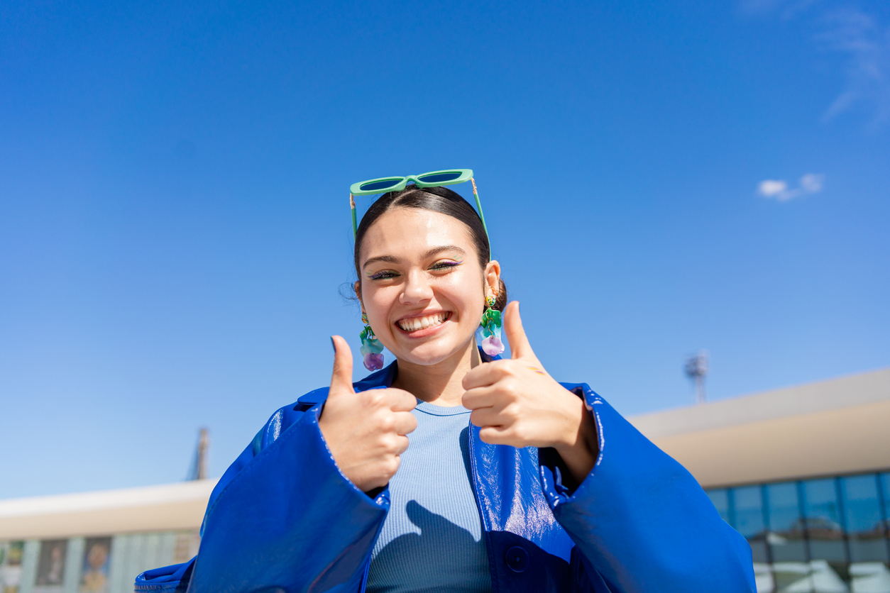 A smiling woman wearing a bright blue jacket and green sunglasses gives two thumbs up outdoors under a clear blue sky.