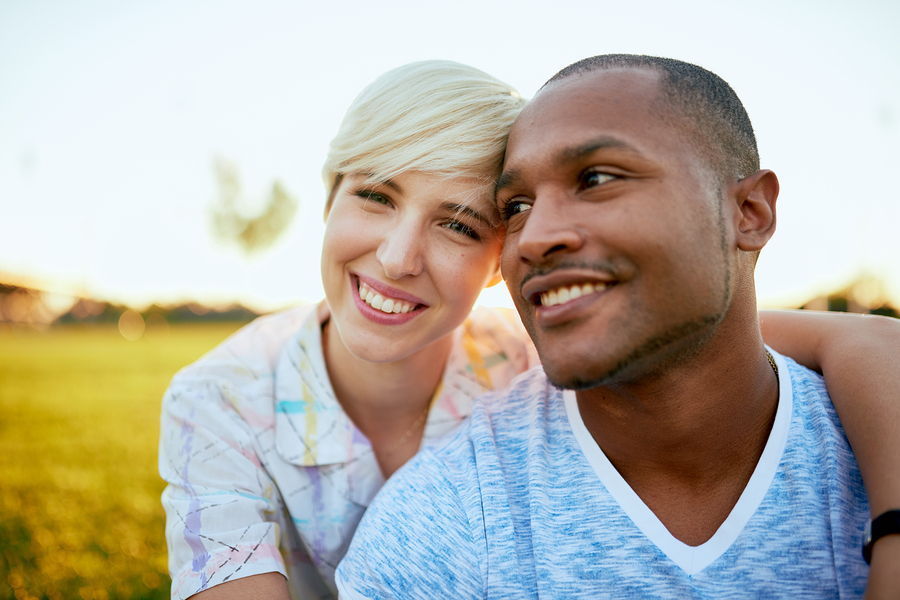 An interracial couple smiles while holding eachother standing in an outdoor setting.