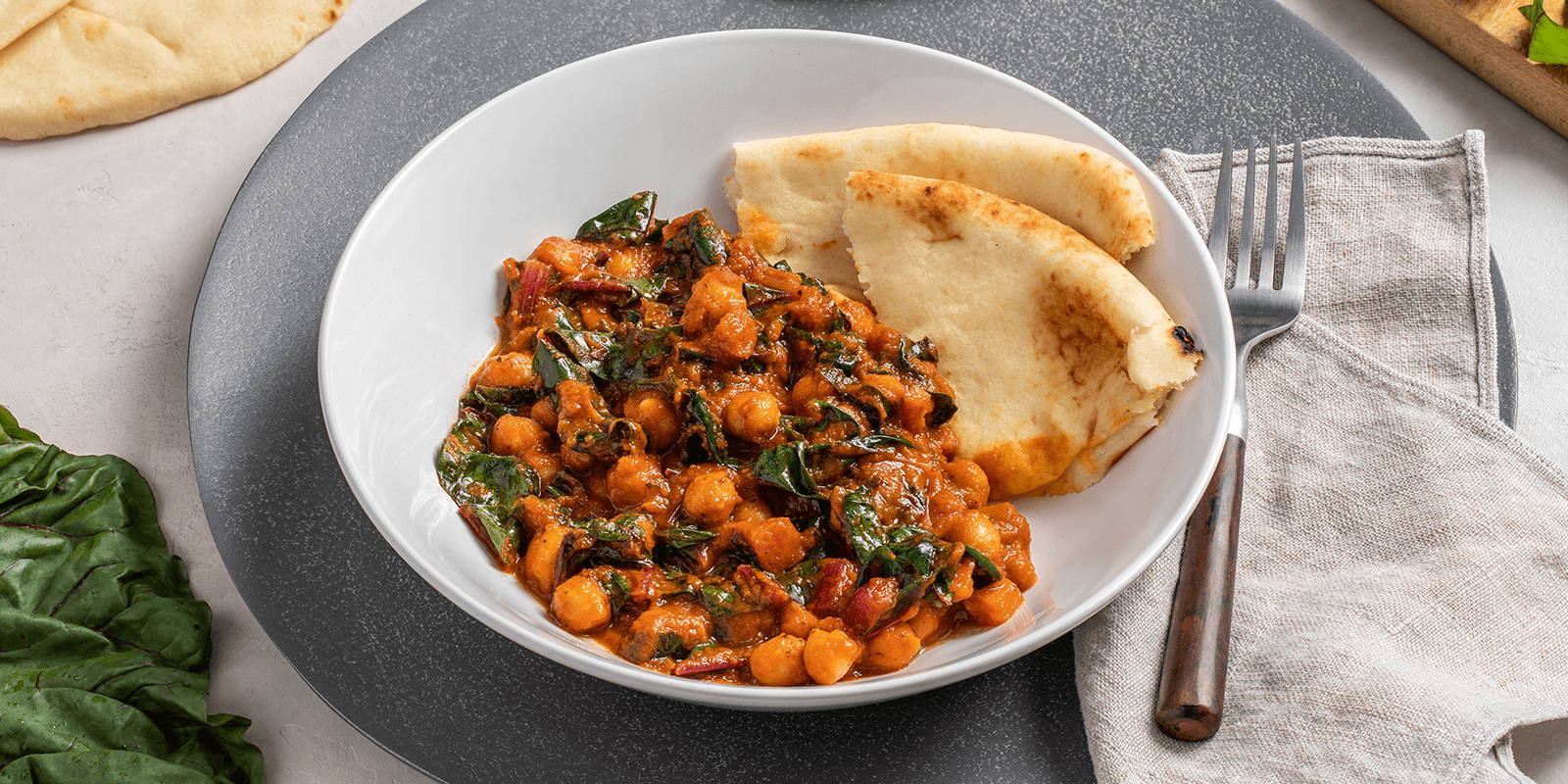 The prepared ZENB Tomato Braised Chickpeas with Swiss Chard recipe, with naan on the side, in a white bowl atop a gray charger plate.