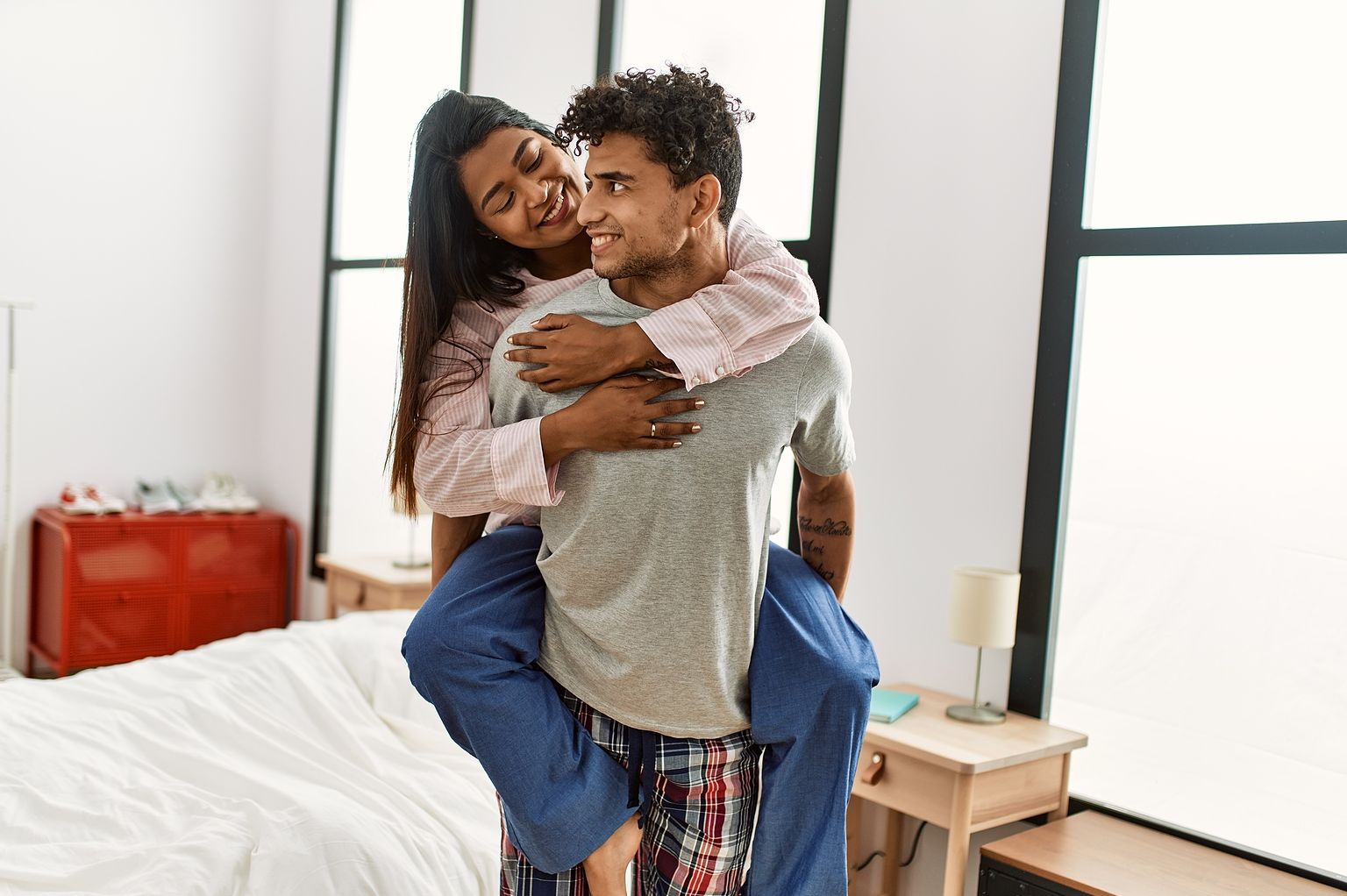 A young latin couple has fun in their room, the man is carrying her while they smile to eachother wearing pijamas.