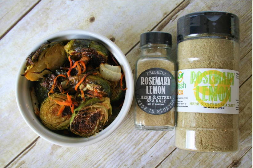 A small white bowl filled with cooked brussels sprouts, onions, and carrots accompanied by a small and large bottle of FreshJax Organic Rosemary Lemon Sea Salt Blend.