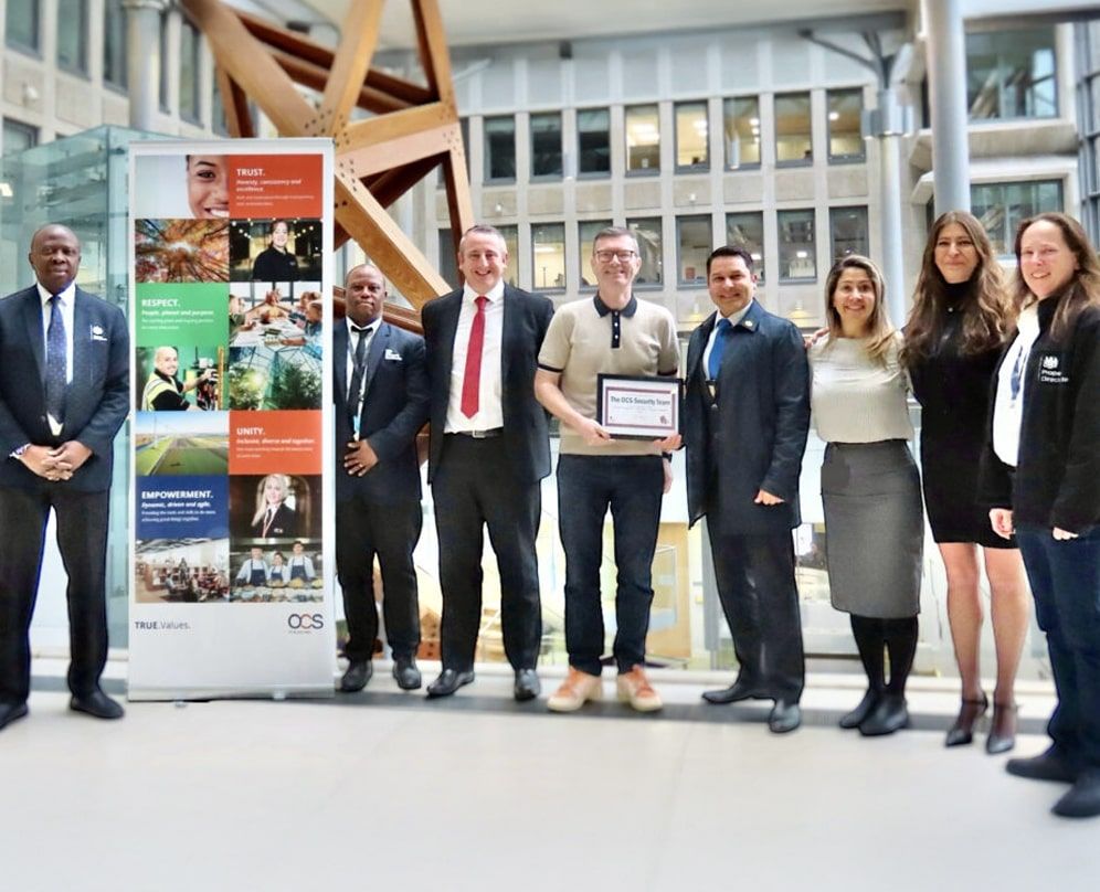A group of eight people stand indoors, some smiling, with one person in the center holding a framed award. Behind them is a colorful banner and a glass structure. The setting appears to be a modern office or corporate space.