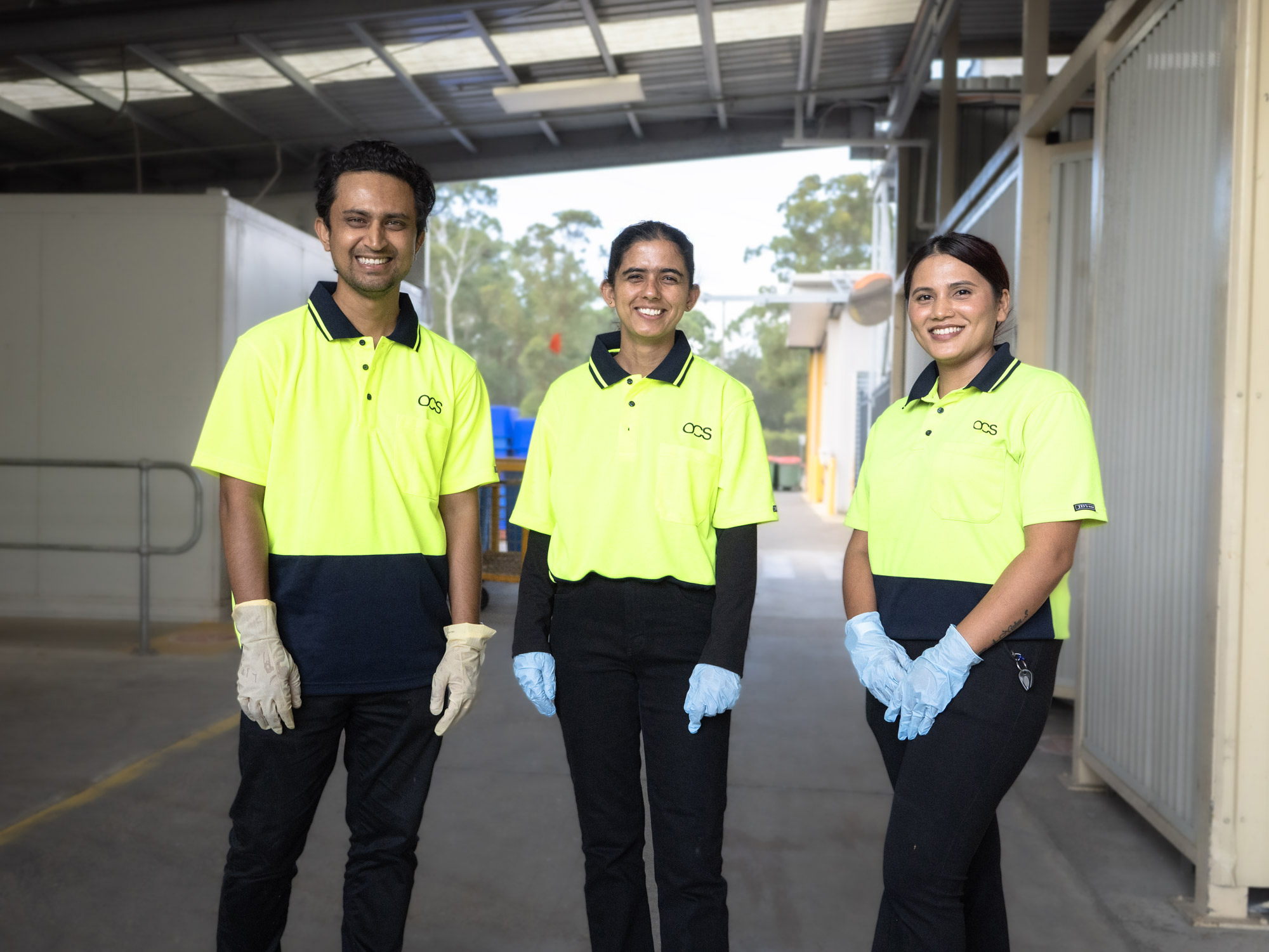 Three people wearing bright yellow and navy uniforms, gloves, and smiling, stand in an industrial or warehouse setting with metal roofing and equipment in the background.