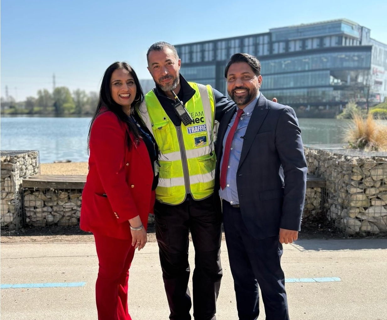 Three people stand together by a lakeside on a sunny day. The person in the middle wears a yellow reflective vest, while the other two, a woman and a man, are dressed in business attire and smiling at the camera.