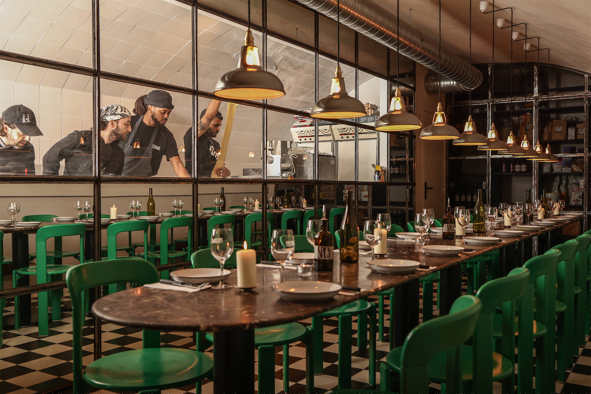 Original Coolicon Antinium 1933 Design lampshades hanging above a long dressed table with green Chairs on either, in front of a pasta kitchen.