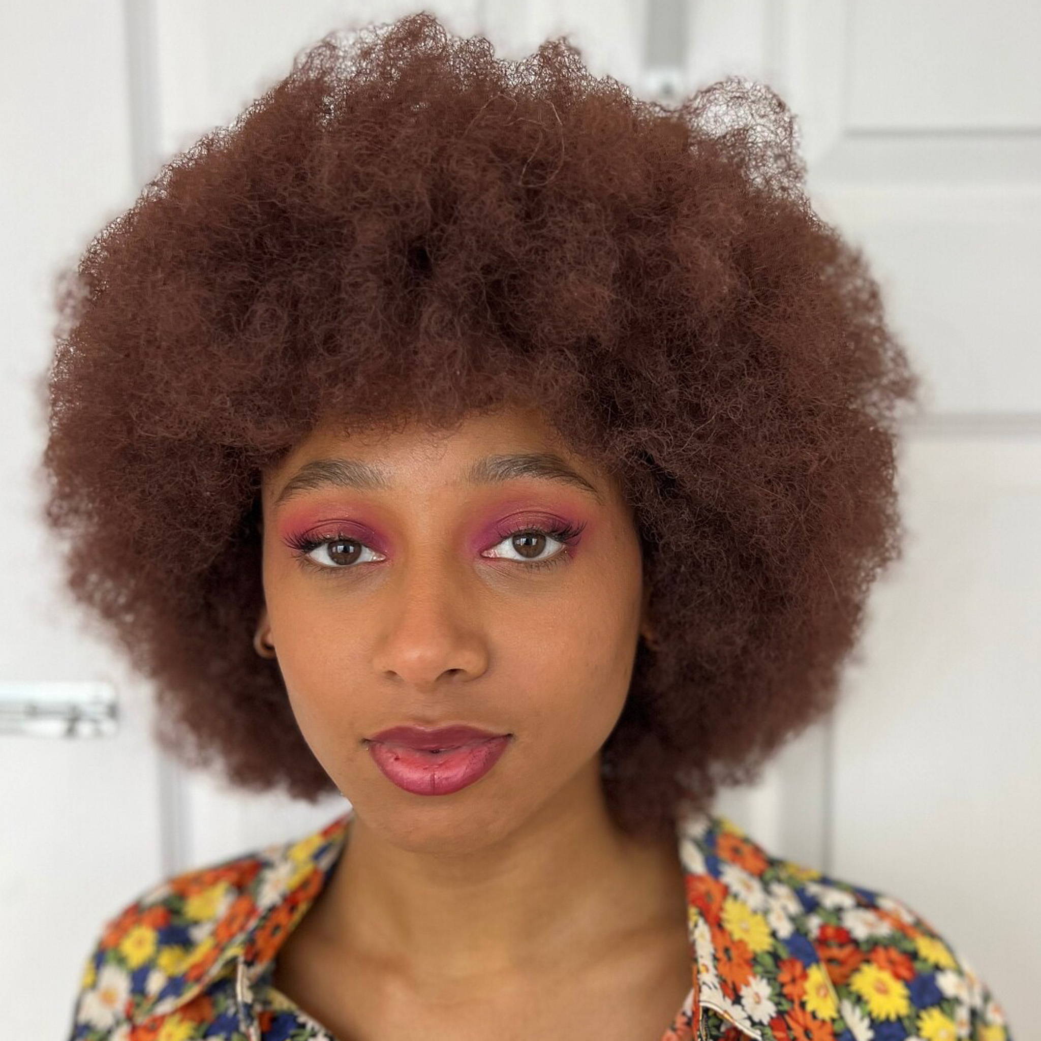 Model Rhianna stands in front of a white door. From the shoulders up, they are wearing a multi-coloured flowery top with a deep purple lipstick and matching eye shadow. 