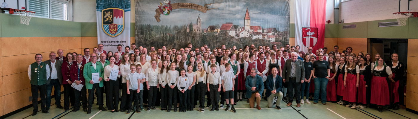 In einer Turnhalle stehen mehrere hundert Menschen in verschiedenen Trachten beieinander und lächeln in die Kamera. 