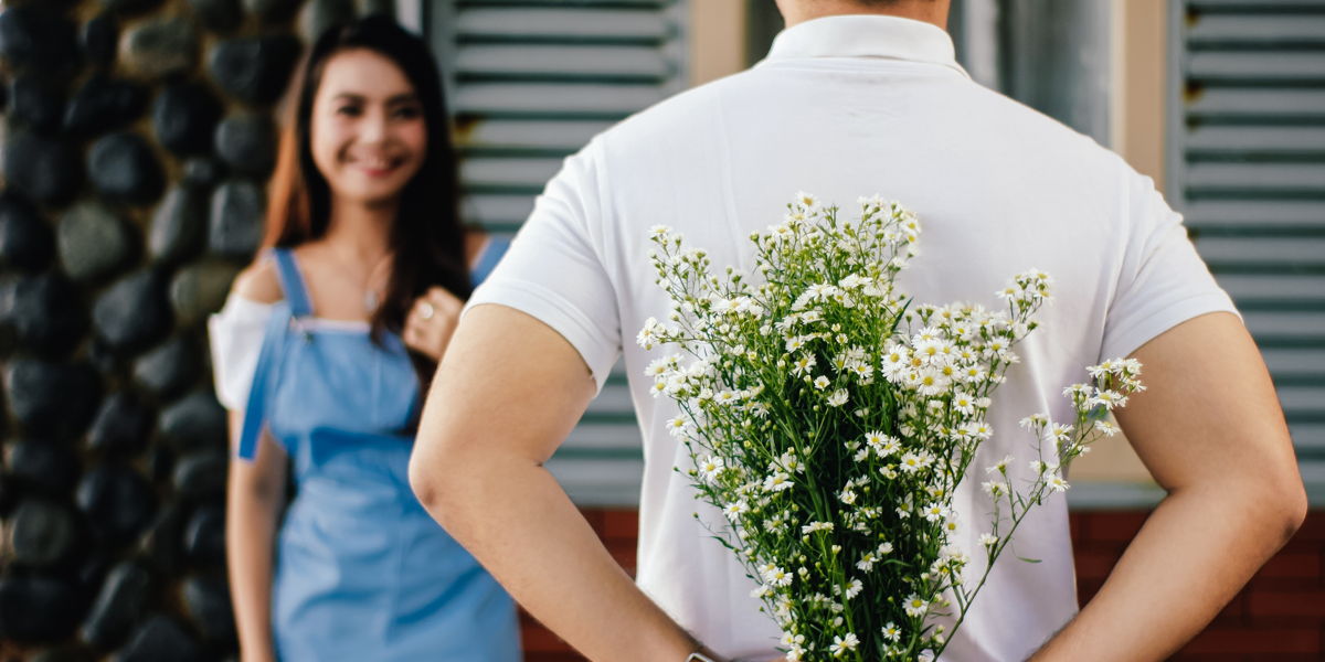 A man holds a bouquet of daisy flowers behind his back ready to surprise a smiling woman.
