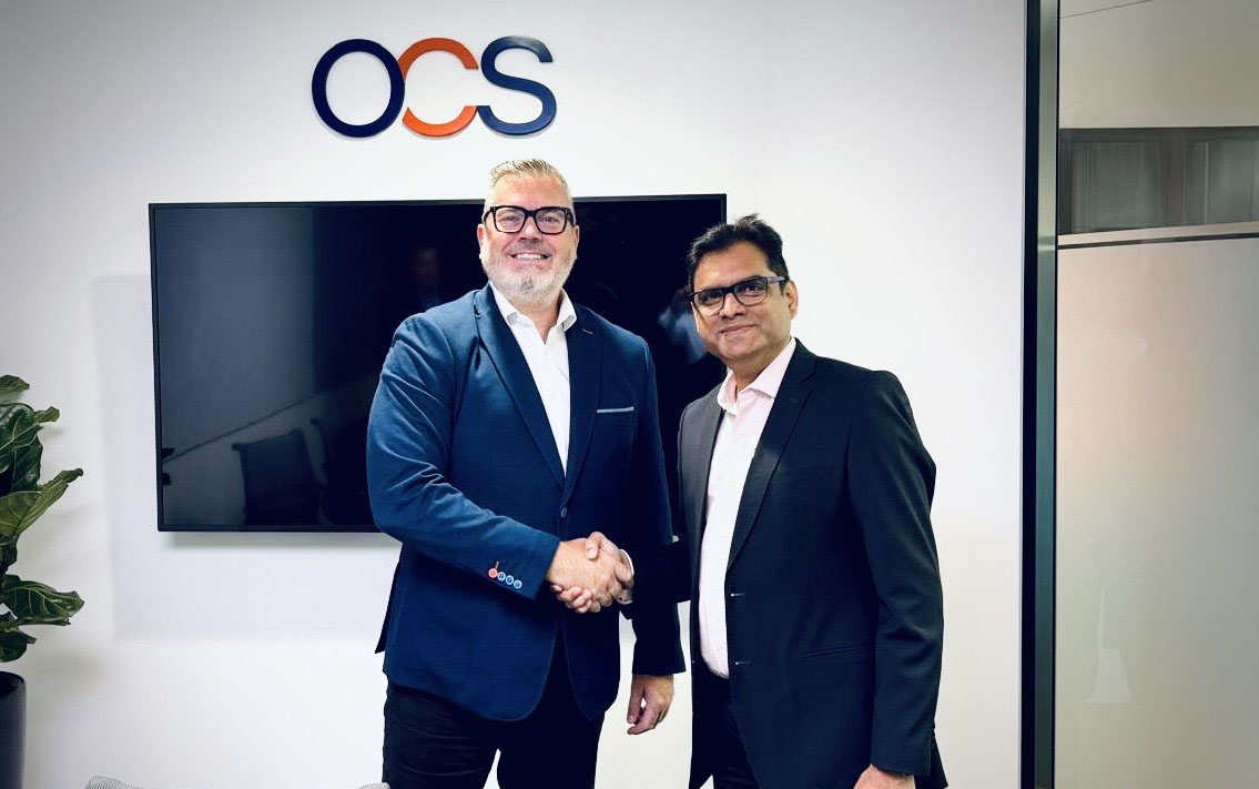 Two men in business attire shake hands and smile in an office setting, standing in front of a TV screen and a wall with the letters OCS displayed above them.