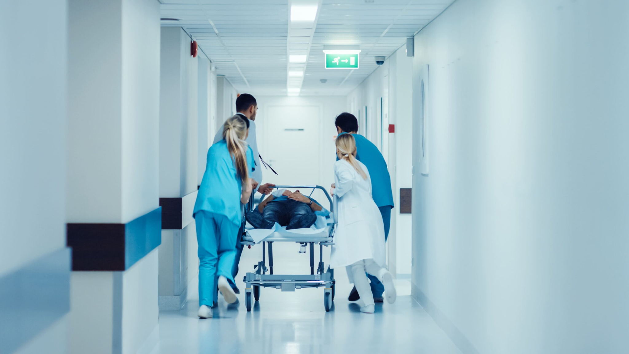 Three medical professionals quickly wheel a patient on a stretcher down a bright hospital hallway, suggesting an emergency situation. The scene is busy and urgent.