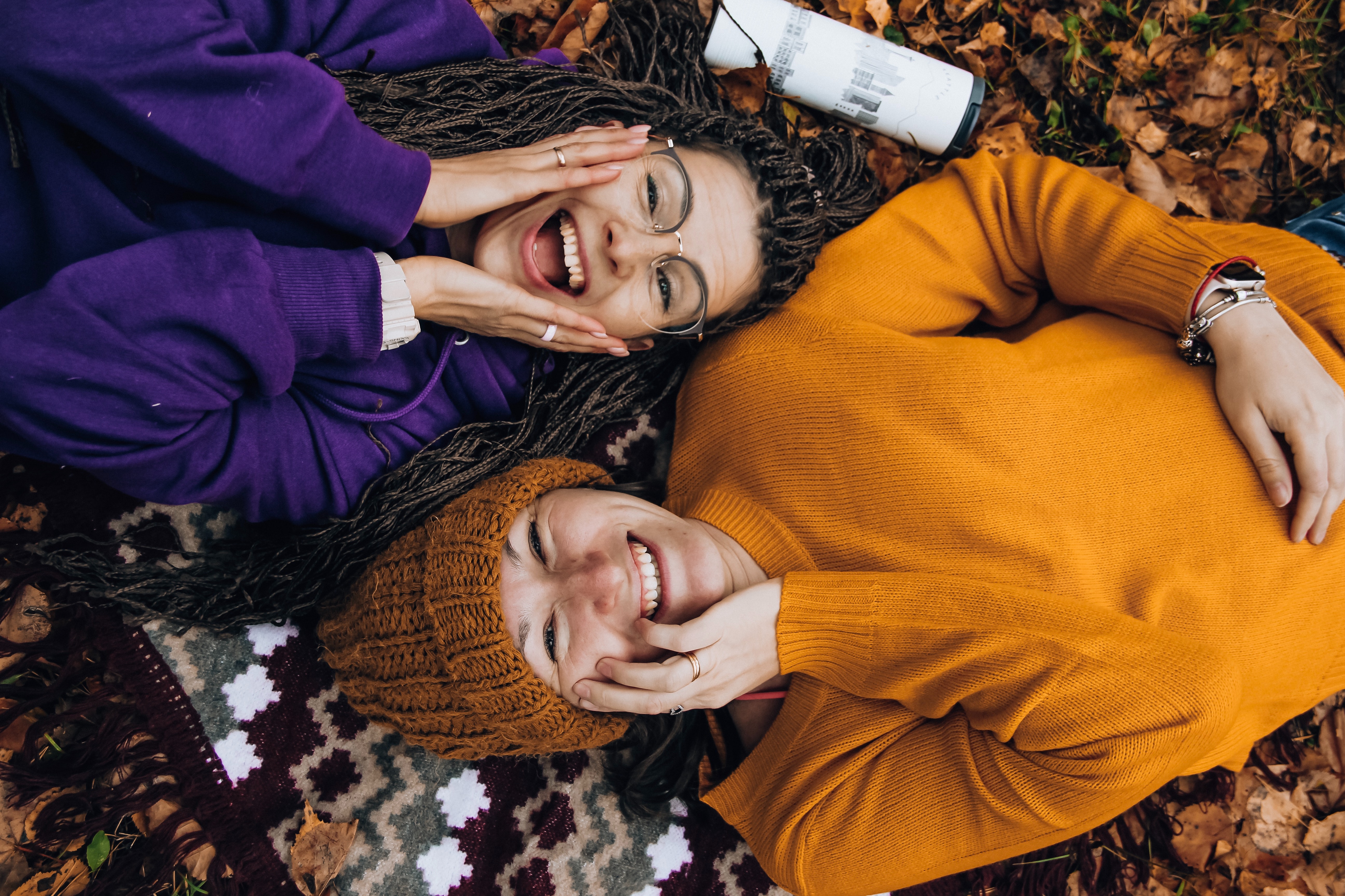 Two women wearing long sleeve sweaters lying on the ground together. Both have a smiling and surprised expression on their faces and are on a blanket.