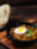 A bowl of stew with a hard-boiled egg and a slice of bread on a wooden cutting board