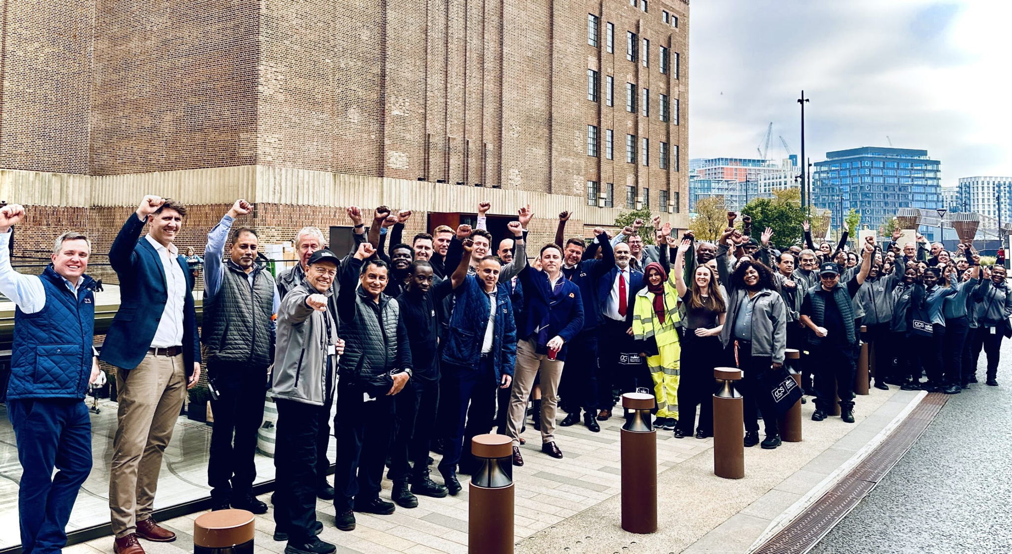 A large, diverse group of people stand outdoors near a brick building, many raising their fists and smiling. Some are in business attire, others wear work uniforms or reflective vests. Urban buildings appear in the background.