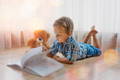 Child reading a book to his teddy bear while lying on the floor.