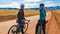 A cyclist is riding a Kensuke gravel bike by KindHuman Bicycles. She is riding outdoors on a paved road in a wheat field. There is fog in the background.