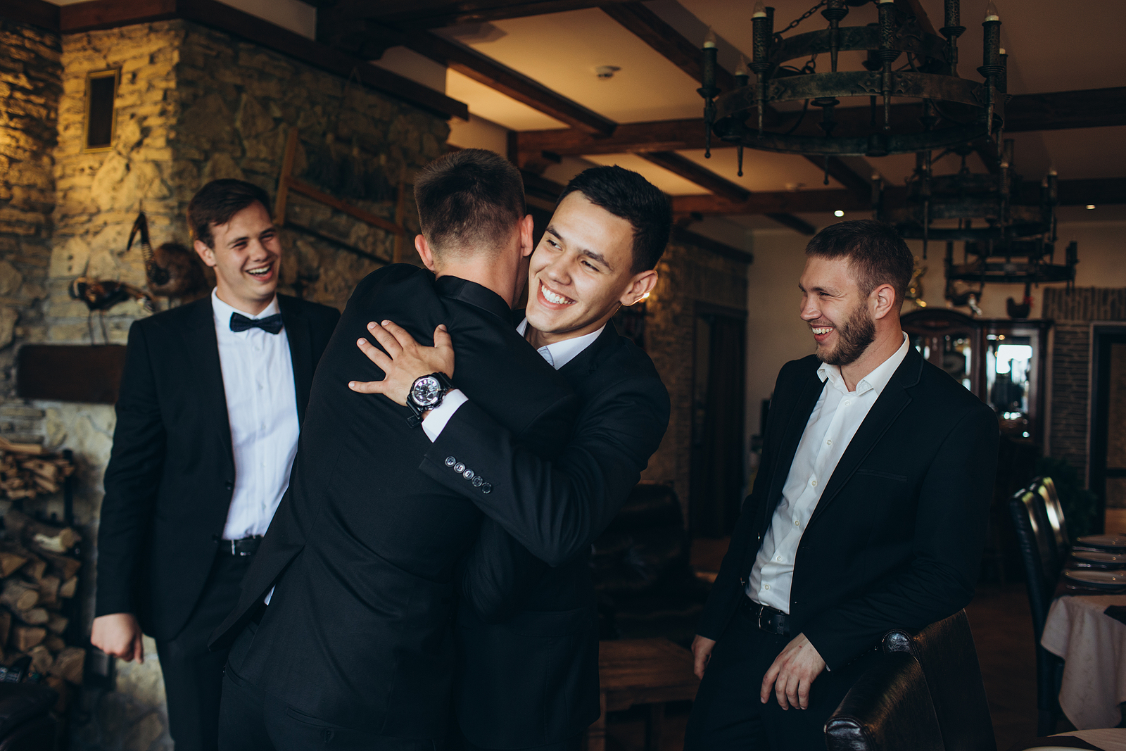 A group of 4 friends wearing tuxedos for a wedding. There are two friends hugging  in the front.